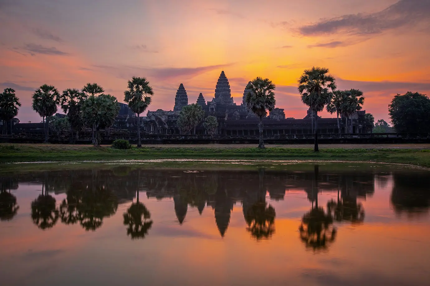 A symmetrical reflection of Angkor Wat and surrounding palm trees across the tranquil moat during a vibrant orange sunrise featured in a travel guide on Cambodia Itinerary.
