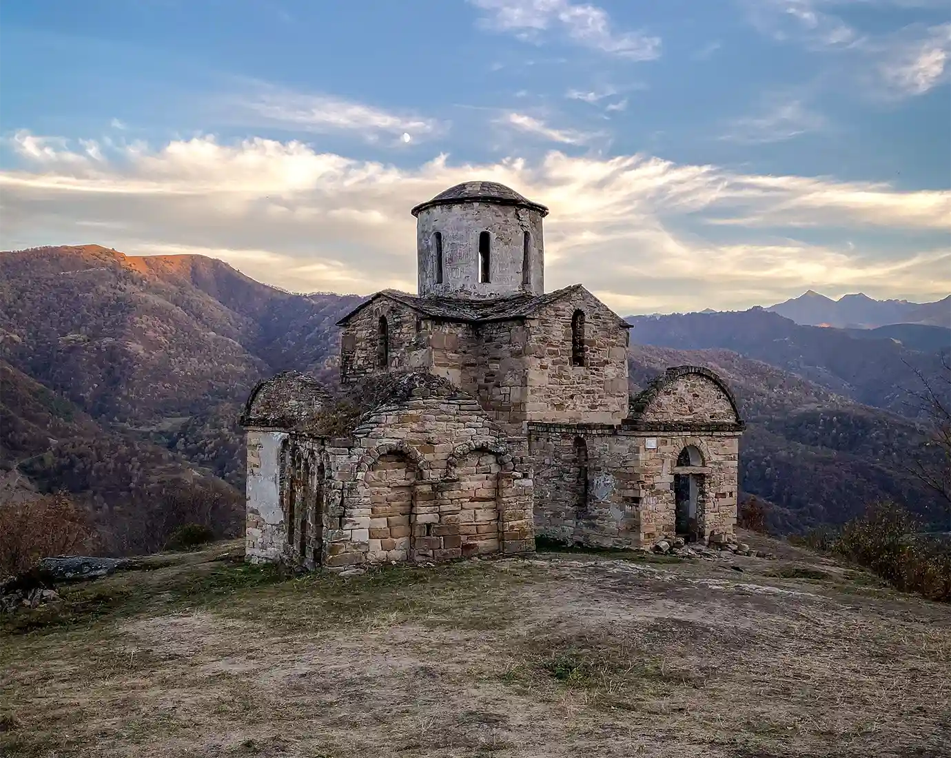 A historic stone church structure standing on a hilltop against a backdrop of distant mountains at sunset, showcasing historical Places to Visit in Georgia.