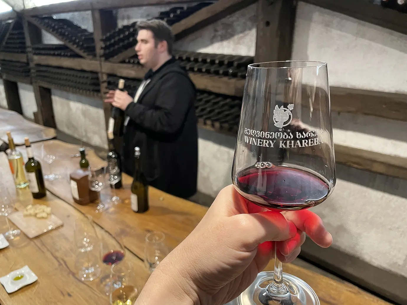 A hand holding a glass of red wine during a tasting tour inside a cellar with bottle racks, illustrating excursion expenses for Georgia Travel Cost.