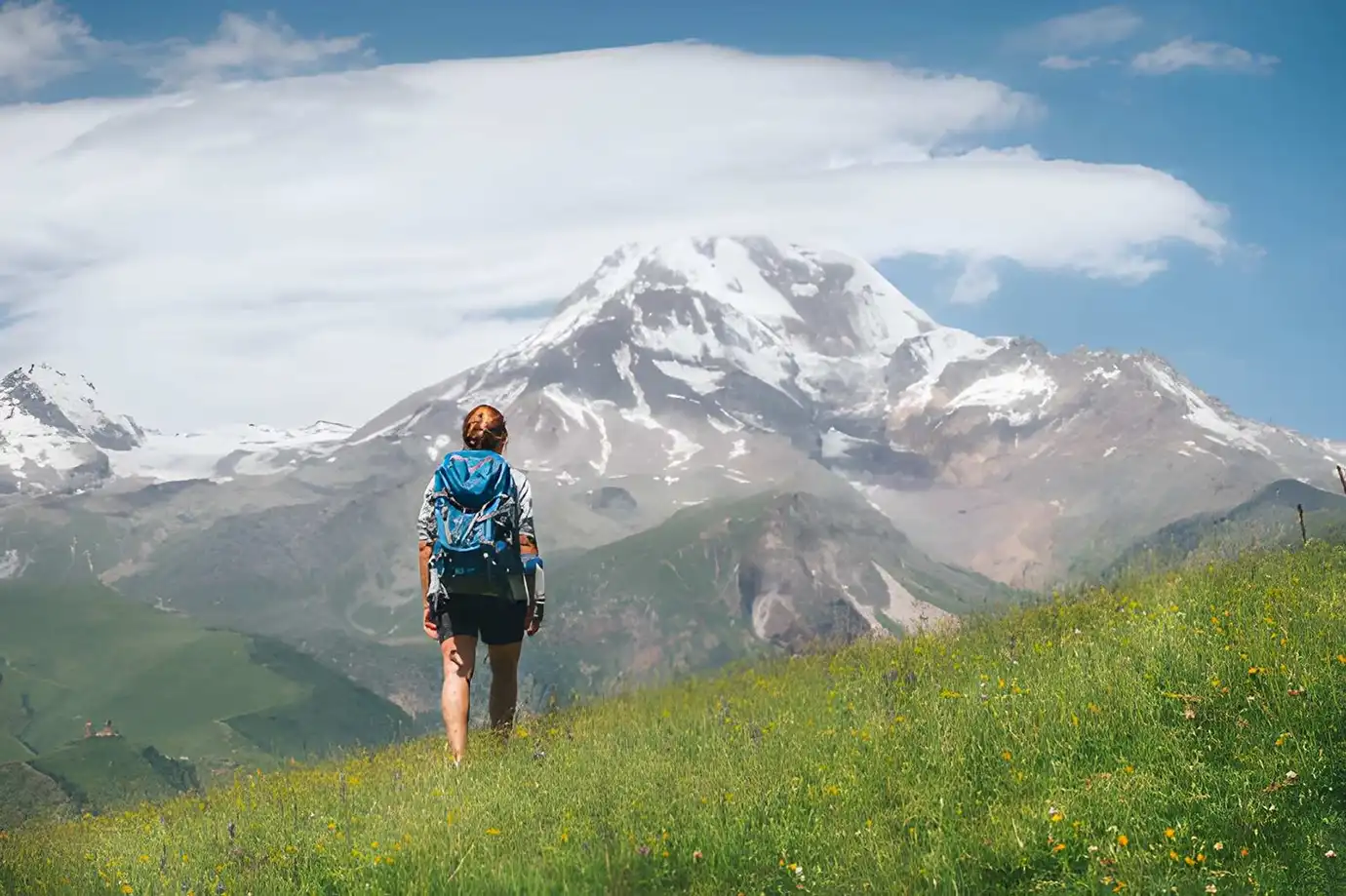 A trekker with a blue backpack walking across a lush green meadow towards snow-capped Mount Kazbek, illustrating ideal hiking conditions for the Best Time to Visit Georgia.