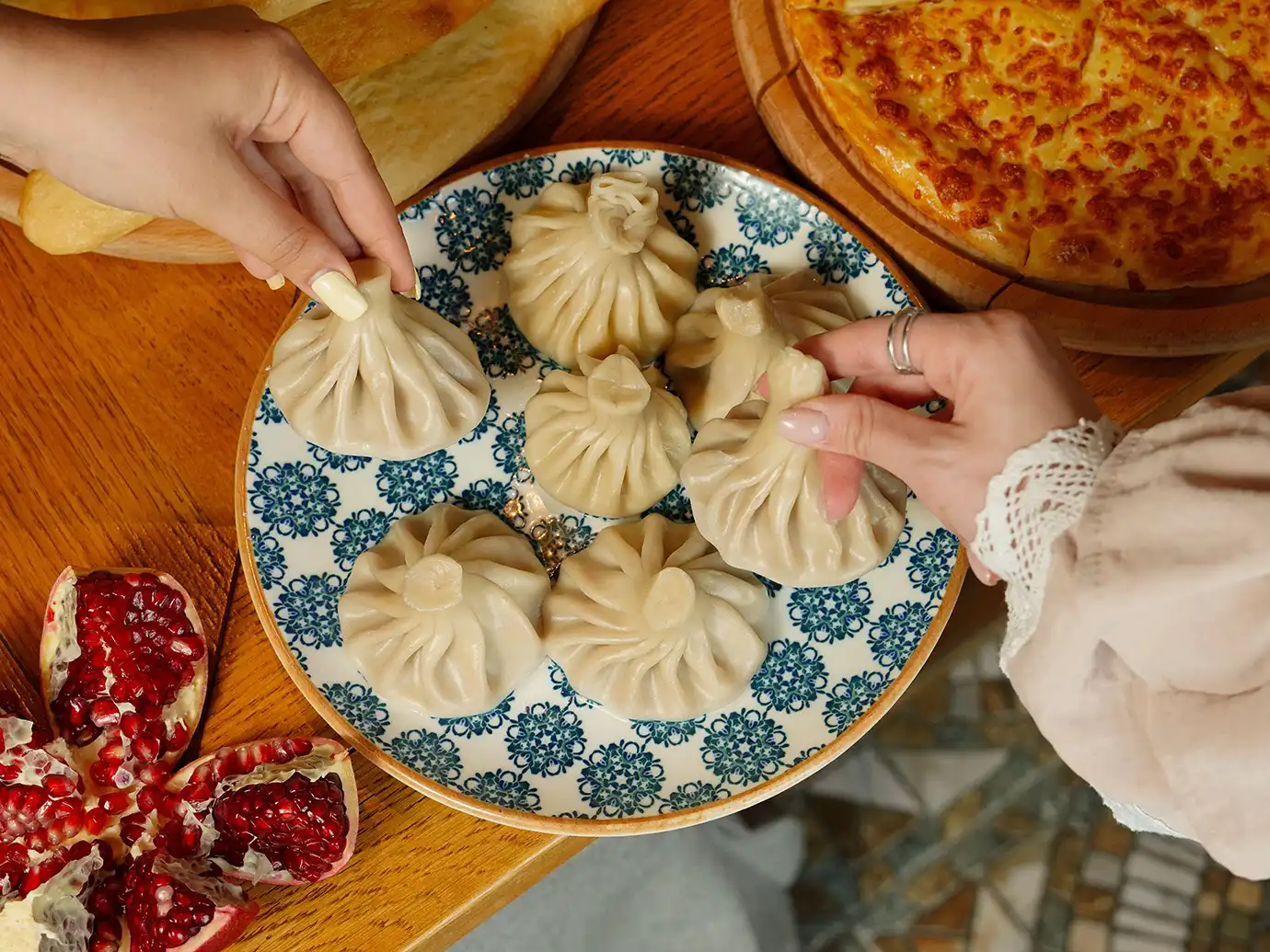 Diners reaching by hand for freshly steamed Khinkali dumplings served on a blue patterned plate, showcasing essential dishes for a Georgian Food Guide.