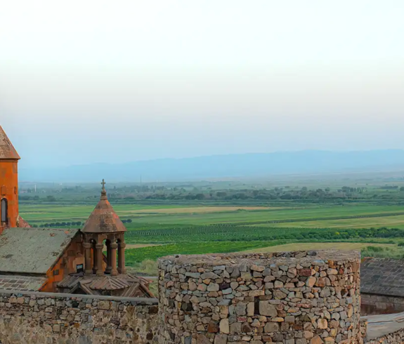 An elevated view of the stone complex of Khor Virap Monastery, featuring red tuff domes and a stone fortification wall overlooking green agricultural fields under a soft dusk sky in Armenia.