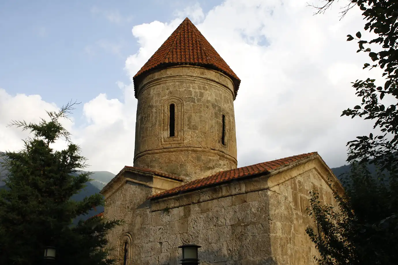 The cylindrical stone tower and red tile roof of the historic Church of Kish surrounded by trees in Kish Village featured in a travel guide on The Ultimate 7-Day Azerbaijan Itinerary.