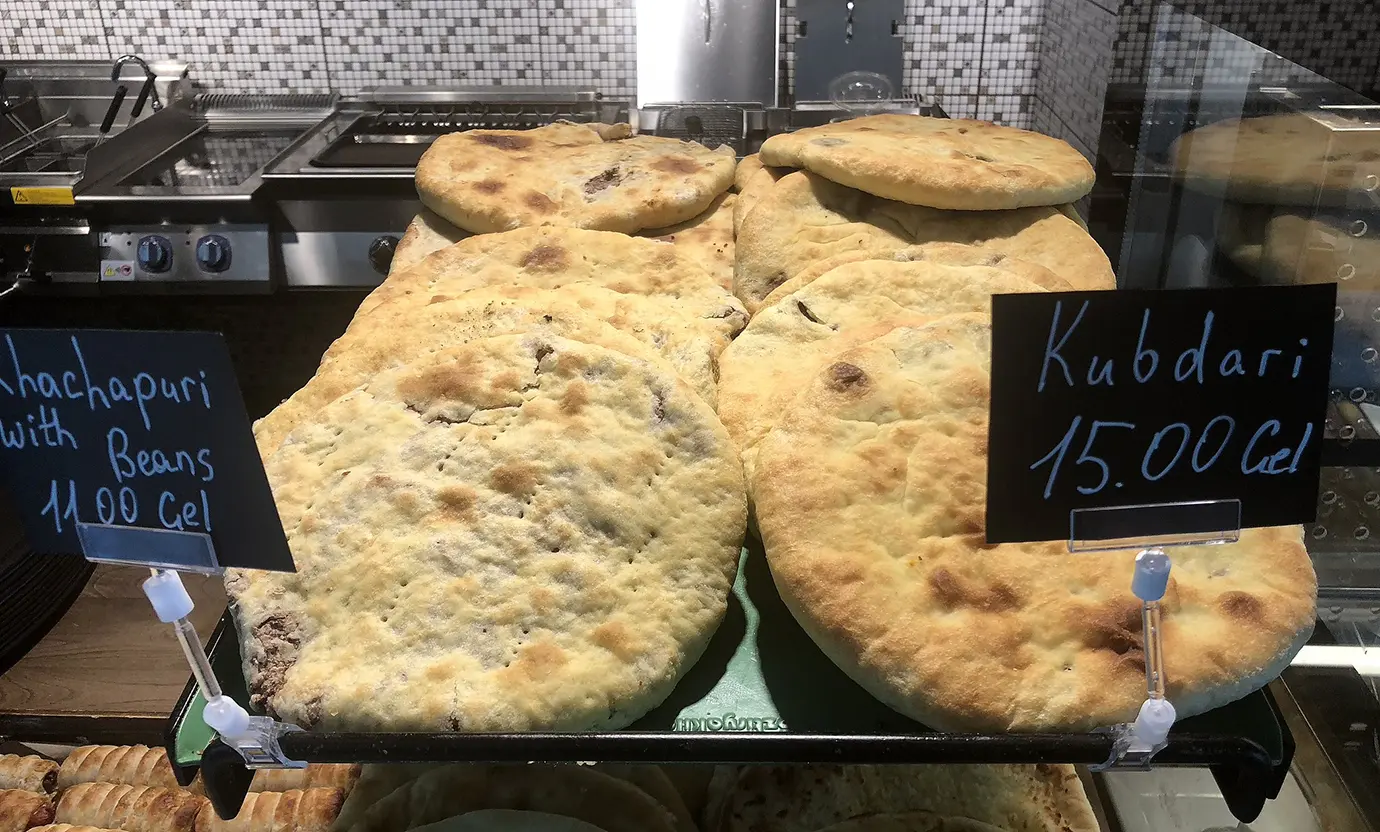 Freshly baked rounds of Svanetian Kubdari meat bread and Lobianibean-filled khachapuri with handwritten price signs on display in a bakery, illustrating recommendations for a Georgian Food Guide.