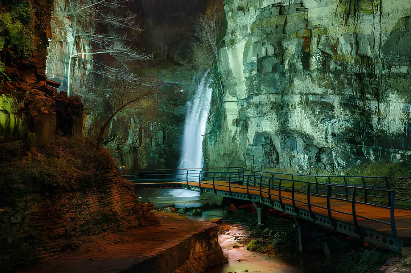 An illuminated night view of the Leghvtakhevi Waterfall flowing down steep canyon cliffs next to a wooden pedestrian walkway, illustrating top natural Things to Do in Tbilisi.