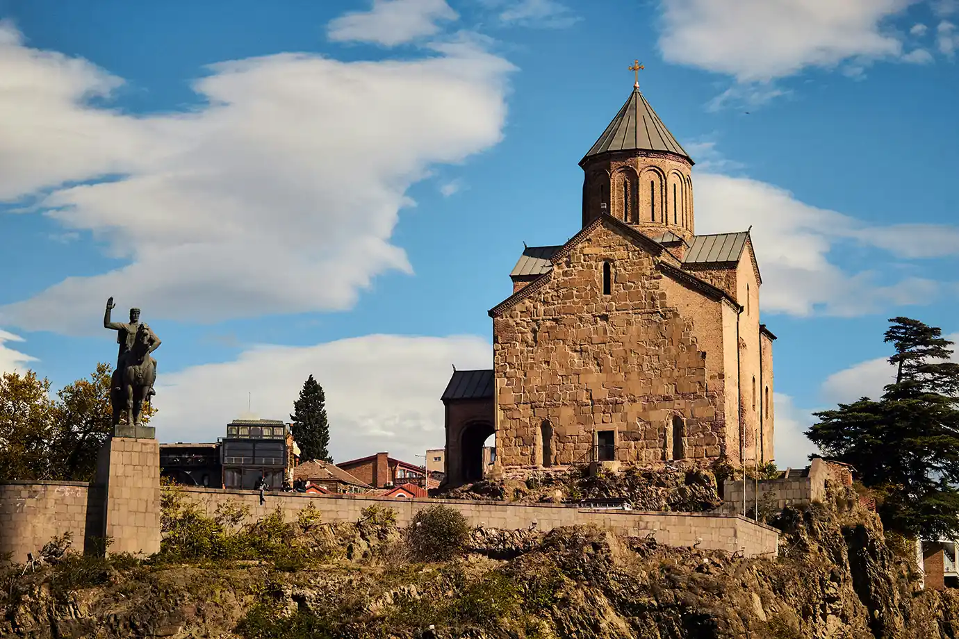 The historic stone Metekhi Church sitting atop a cliff beside the equestrian monument of King Vakhtang Gorgasali under a blue sky, showcasing iconic Things to Do in Tbilisi.