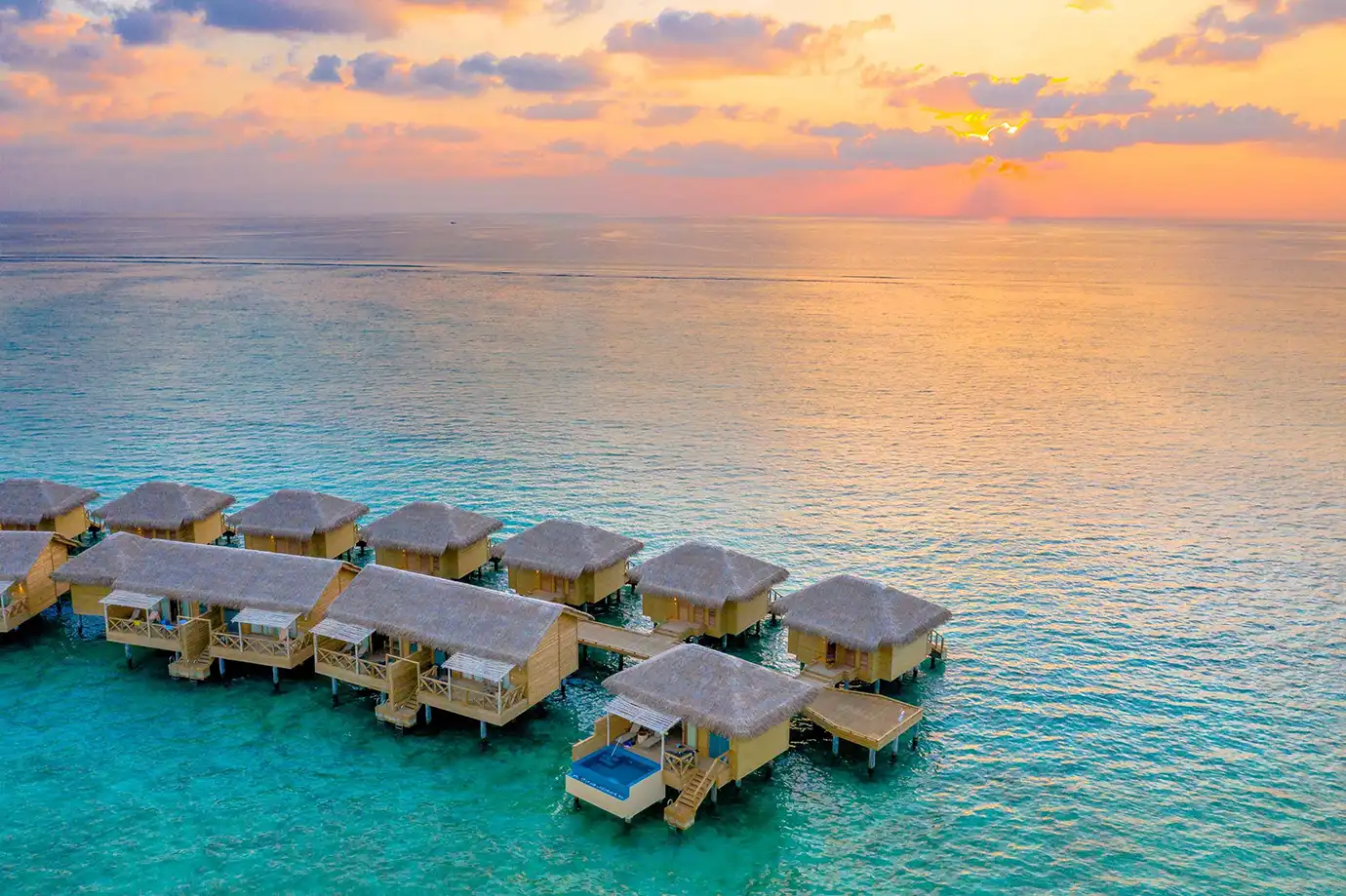 A line of thatched-roof overwater villas stretching into clear turquoise waters at sunset featured in a travel guide on the Best Time to Visit the Maldives.