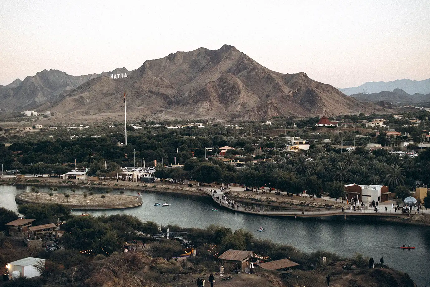 An elevated wide shot of a winding lake with a pedestrian bridge and small boats, framed by green trees, a small island, and rugged Hajar mountains featuring the white HATTA sign under a soft dusk sky.