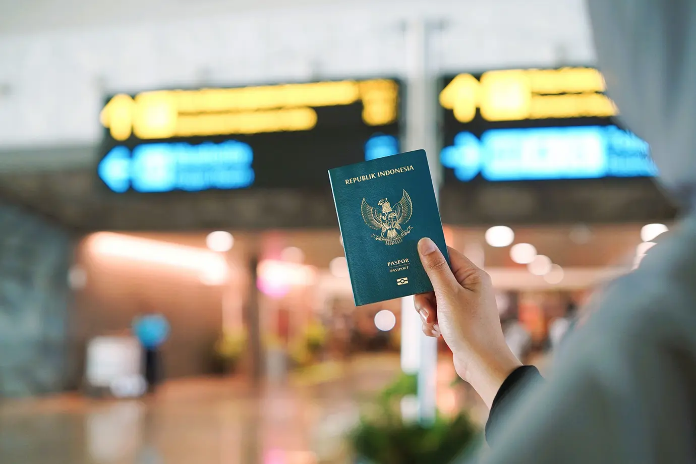 A traveler holding a green Indonesian passport inside an airport terminal with blurred directional flight signs in the background, essential for understanding Dubai Visa Requirements.