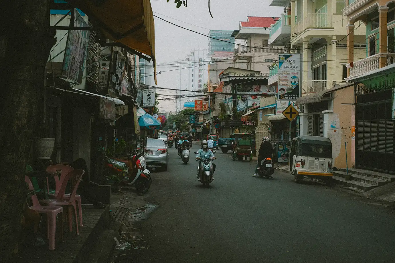 Motorbikes and a tuk-tuk navigating a urban street lined with multi-story buildings and shops featured in a travel guide on Is Cambodia Safe.