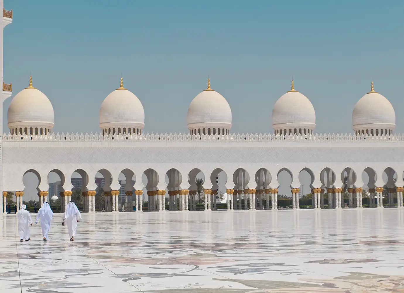 Three men in traditional white kanduras walking across the reflective marble courtyard of Sheikh Zayed Grand Mosque, framed by ornate white arcades and five white domes with golden finials under a clear blue sky in Abu Dhabi.