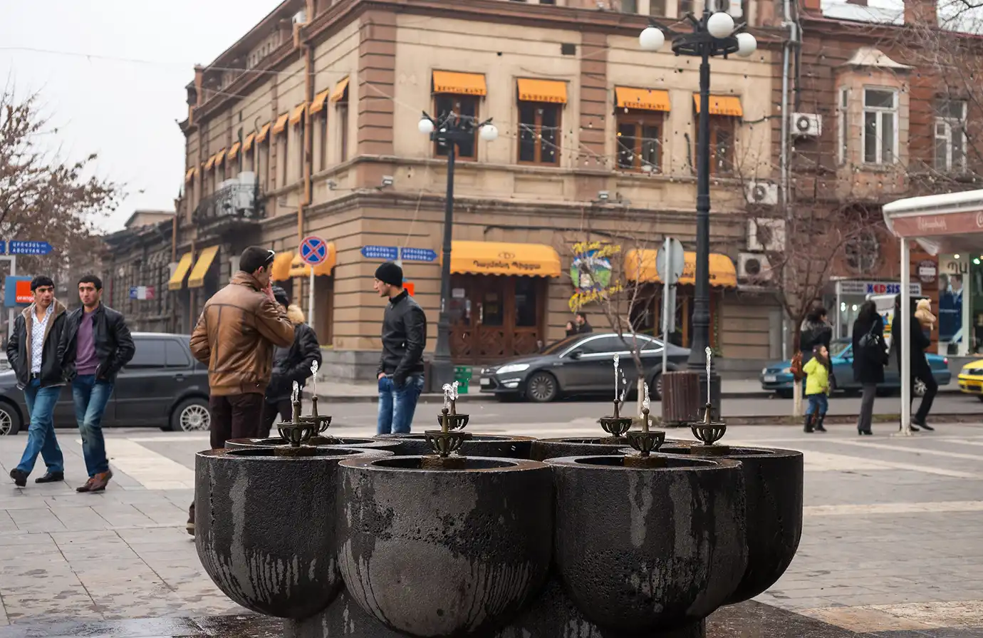 A multi-spouted stone pulpulak drinking water fountain in Yerevan with street traffic and classical stone architecture in the background, featured in a travel guide on Best Things to Do in Yerevan Armenia.