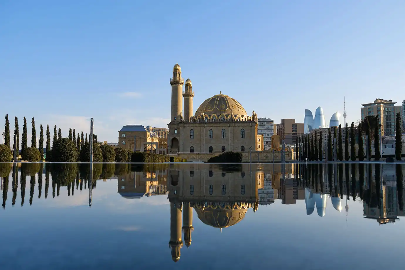 A wide landscape view of the historic Taza Pir Mosque and surrounding cypress trees perfectly reflected on a large, still pool of water, with the modern Flame Towers rising in the distance under a clear blue sky in Baku, Azerbaijan.