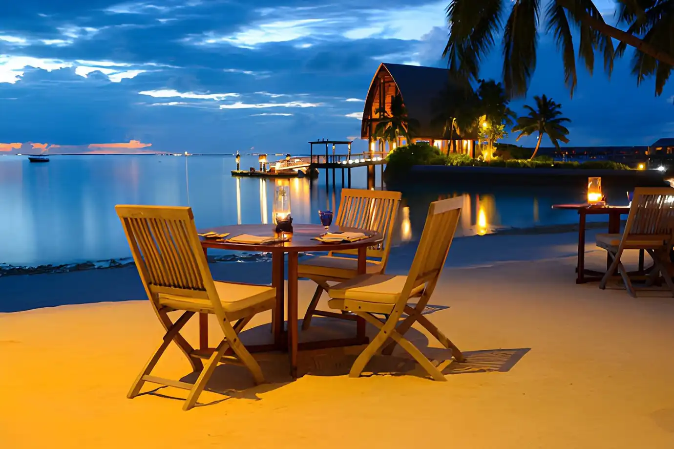 A candlelit wooden dining table set on a white sand beach at dusk near a seaside resort featured in an article asking Is a Maldives All-Inclusive Luxury Resort Holiday Worth It?