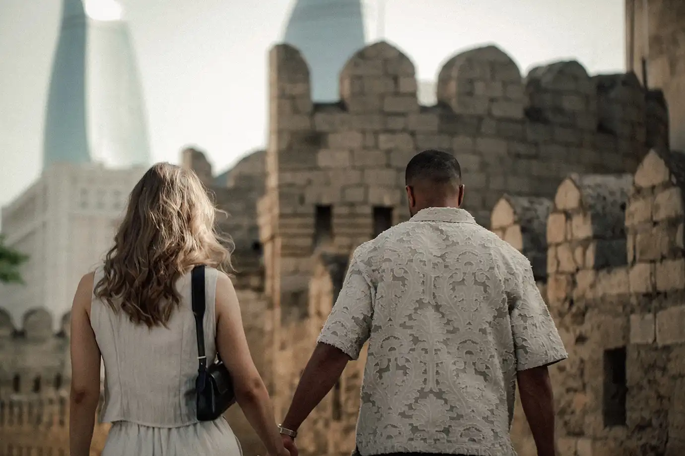 A couple holding hands seen from behind as they walk along the stone fortress walls of Old City Baku, with the modern, out-of-focus Flame Towers rising in the soft-lit background.
