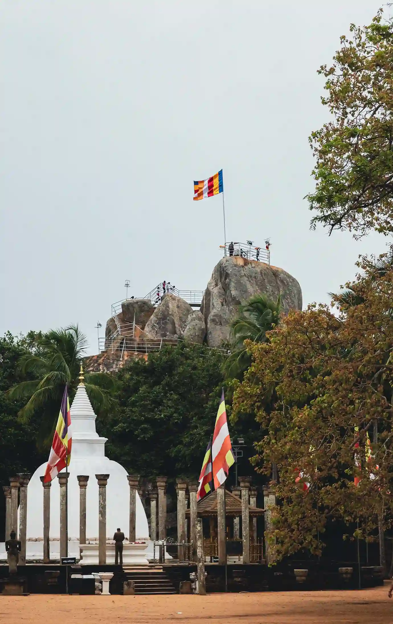 White stupa surrounded by stone pillars with the Aradhana Gala rock peak and Buddhist flags fluttering at Mihintale.