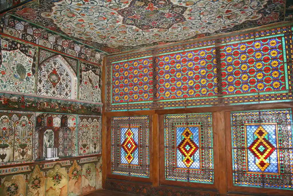 Vibrant red, blue, and yellow geometric stained-glass Shebeke windows illuminate hand-painted floral wall frescoes inside the Palace of Sheki Khans featured in a travel guide on The Ultimate 7-Day Azerbaijan Itinerary.