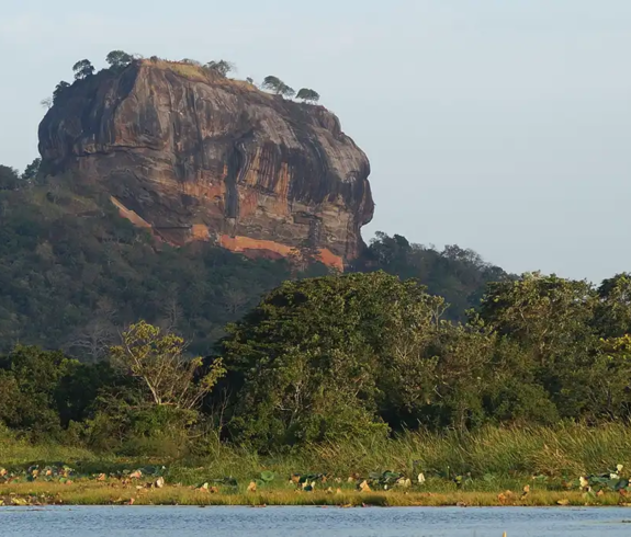 A wide landscape photograph showing the massive granite monolith of Sigiriya Rock Fortress rising above green forest canopy and a calm lake at morning light in Essence of Sri Lanka