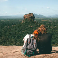 A wide high-angle photograph shot from behind showing a traveling couple sitting on a rocky peak, taking in the panoramic landscape view of Sigiriya Rock Fortress rising above green jungle plains in Sri Lanka. Essence of Sri Lanka