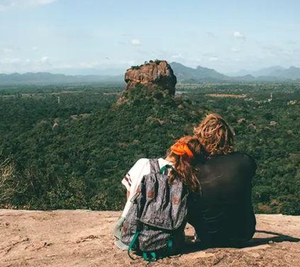 A wide high-angle photograph shot from behind showing a traveling couple sitting on a rocky peak, taking in the panoramic landscape view of Sigiriya Rock Fortress rising above green jungle plains in Sri Lanka. Essence of Sri Lanka
