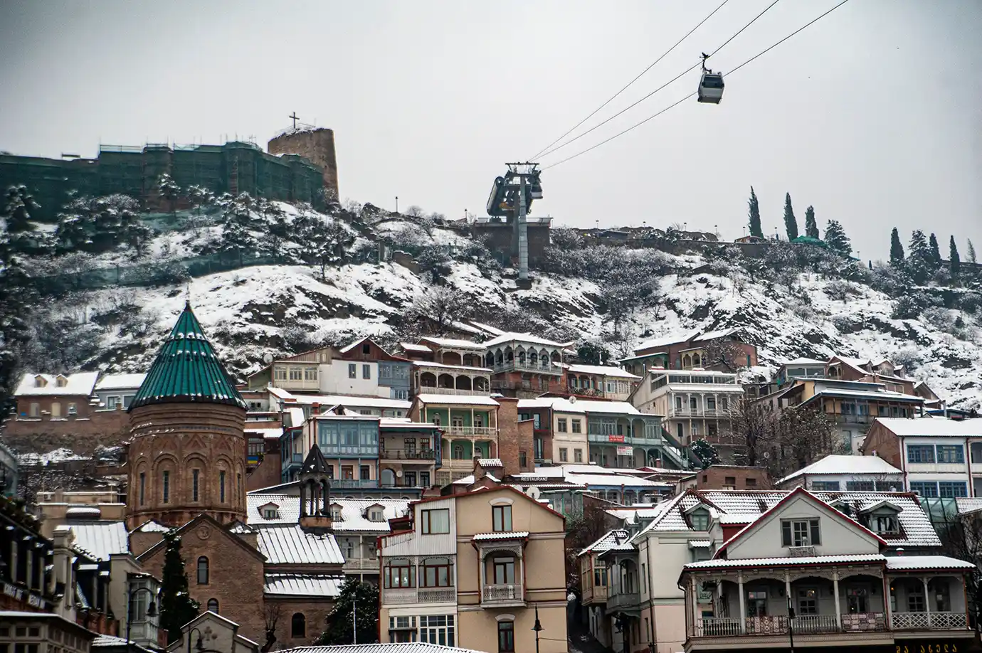 A winter landscape of Old Tbilisi showing snow-covered rooftops, Narikala Fortress on the hill, and an aerial cable car, representing the Best Time to Visit Georgia.