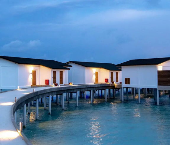 A curving white boardwalk lit by modern warm lights leading to overwater bungalows at dusk featured in a guide on How to Plan a Maldives Luxury Honeymoon on an Affordable Budget.