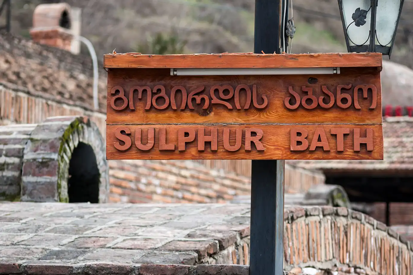 A wooden sign reading "SULPHUR BATH" in English and Georgian script set in front of the brick domed bathhouses of Abanotubani, showcasing iconic Things to Do in Tbilisi.