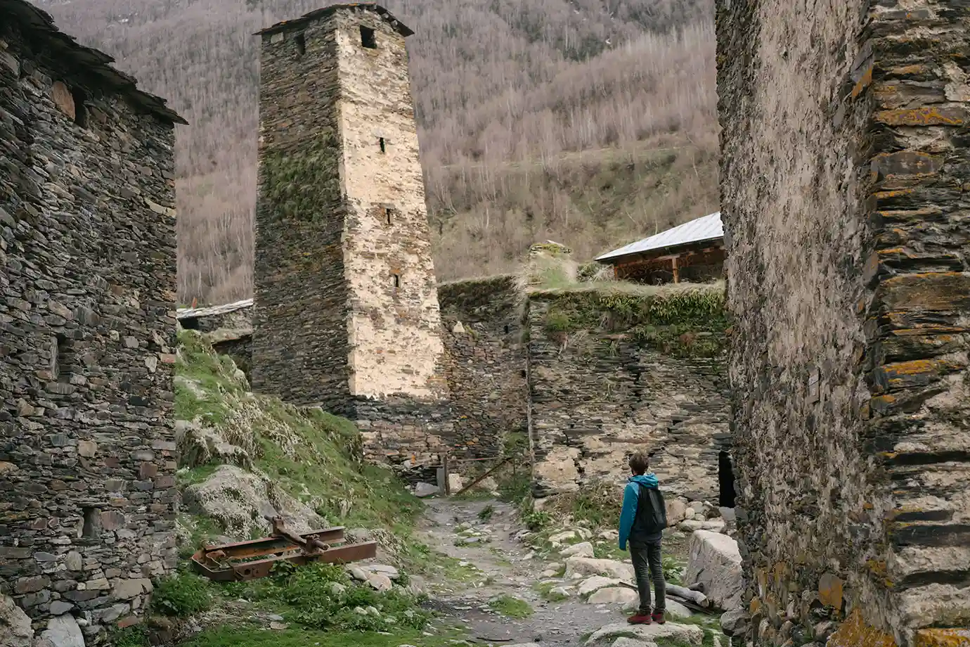 A lone hiker standing in a narrow stone path surrounded by ancient stone houses and a tall Svan tower, highlighting cultural exploration during the Best Time to Visit Georgia.
