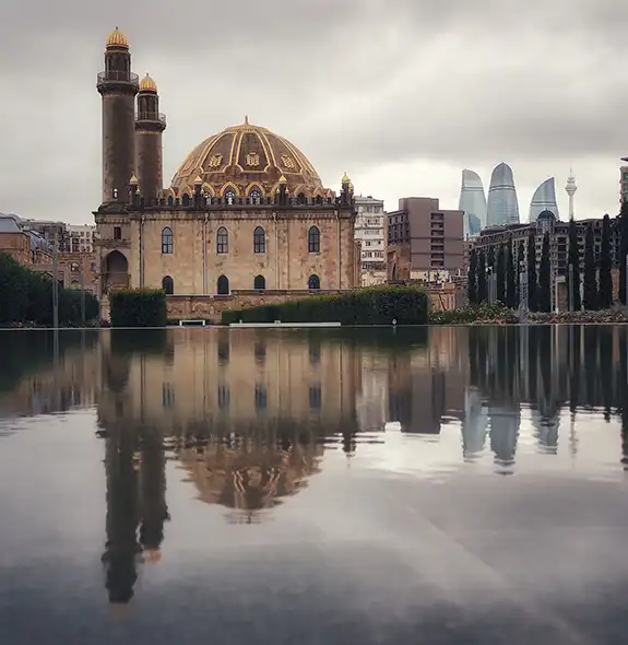 The stone structure, golden dome, and twin minarets of Taza Pir Mosque reflecting symmetrically on a calm pool of water, with the modern Flame Towers visible in the background under an overcast sky in Baku, Azerbaijan.