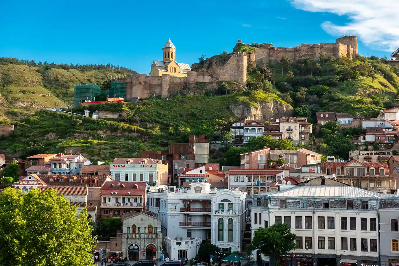 A daylight view of colorful historic buildings in Old Tbilisi with Narikala Fortress perched on the hillside above, representing classic Places to Visit in Georgia.