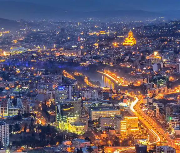 A panoramic night view of the glowing Tbilisi city skyline with illuminated bridges, modern buildings, and Holy Trinity Cathedral, highlighting top Things to Do in Tbilisi.