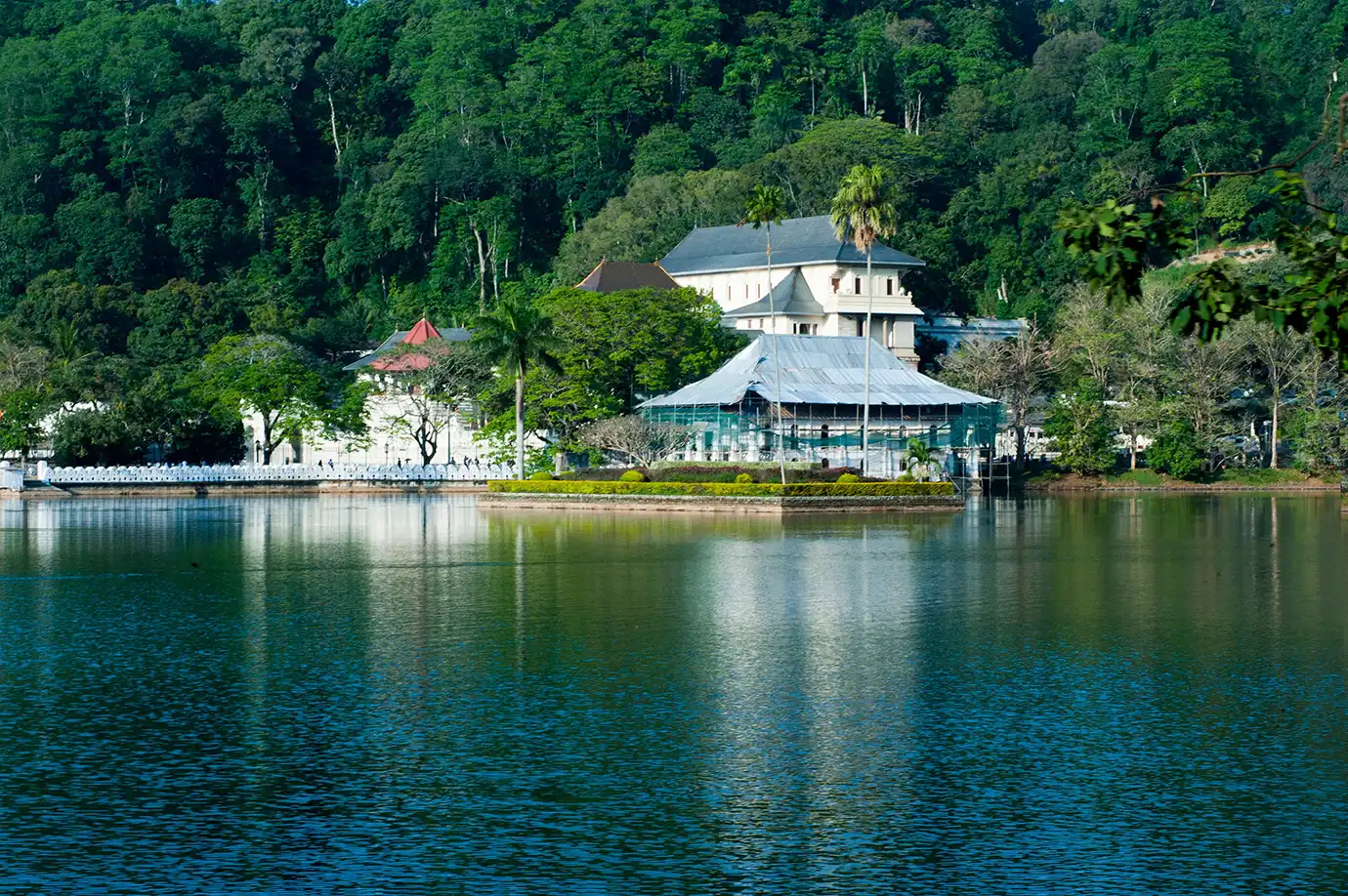 A wide landscape photograph showing the historic Temple of the Tooth complex and Sri Dalada Maligawa reflected in the calm waters of Kandy Lake, backed by dense green forested hills in Kandy, Sri Lanka.