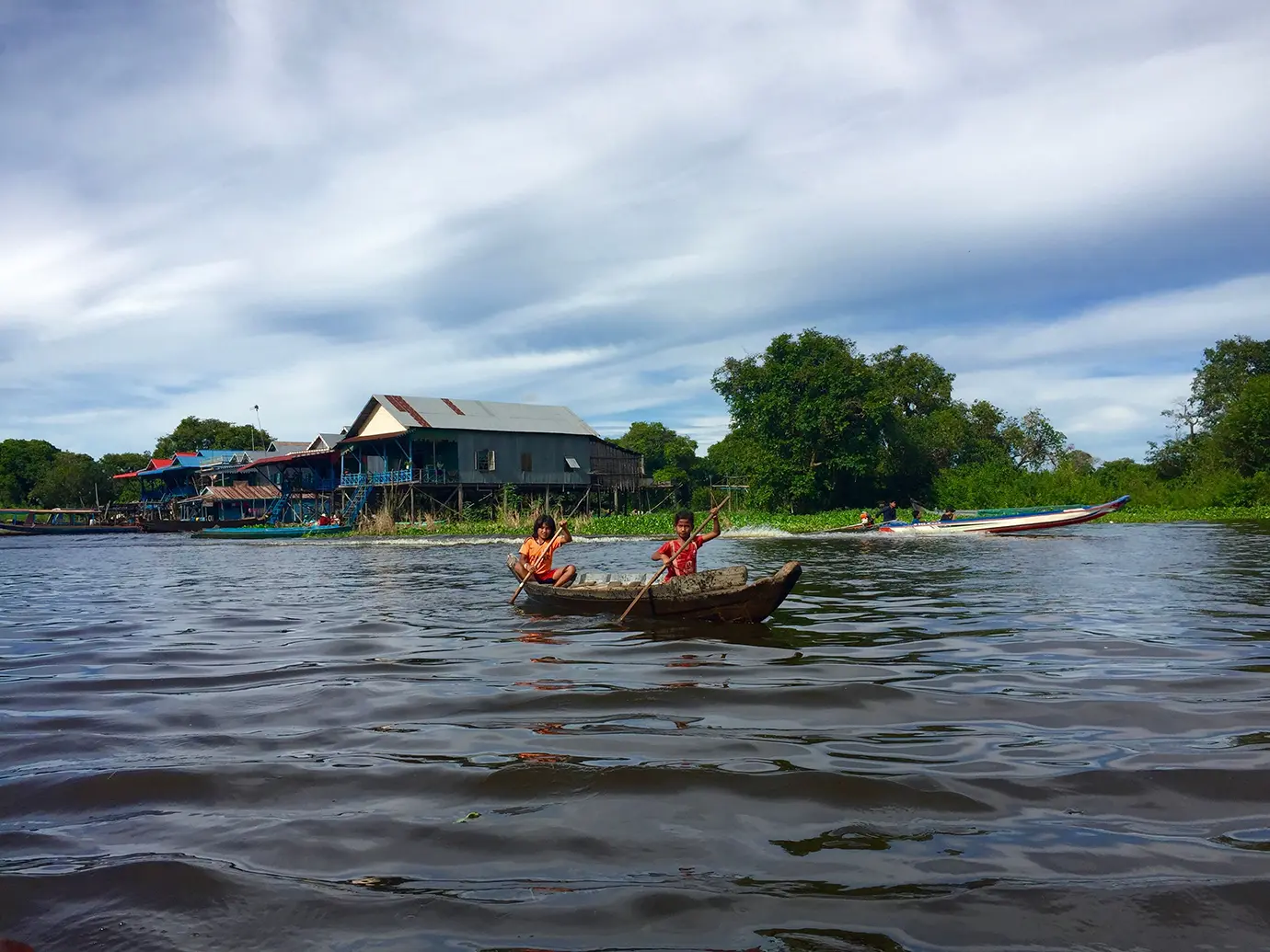Two local Cambodian children rowing a small wooden boat through high water past stilted wooden houses in a floating village on Tonle Sap Lake featured in a travel guide on the Best Time to Visit Cambodia.