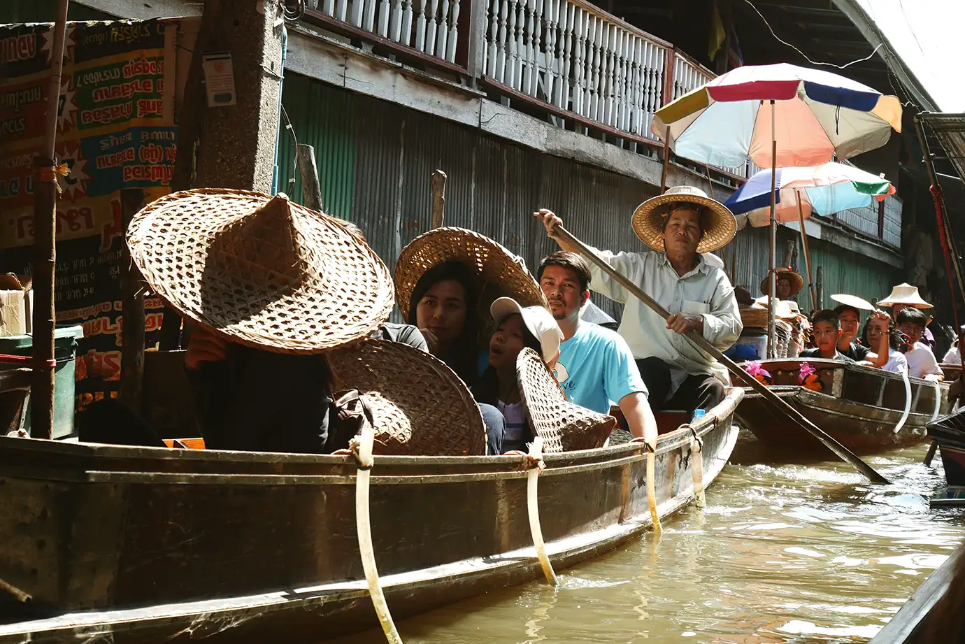 A close-up high-angle photograph of passengers wearing conical straw hats riding in a long wooden sampan boat steered by a boatman down a narrow canal in Bangkok, Thailand.