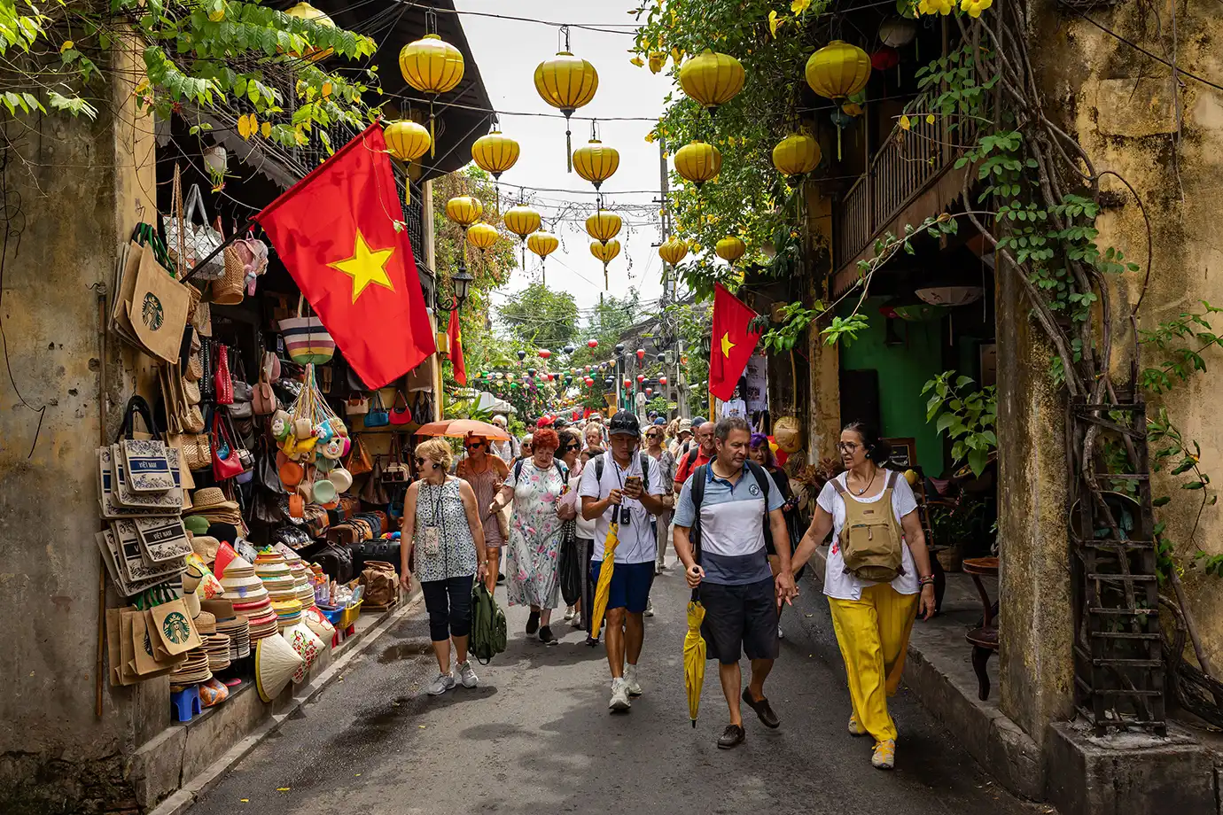 A wide eye-level photograph of tourists walking along a narrow paved street flanked by yellow historic shophouses, hanging yellow lanterns, and red Vietnamese flags in Hoi An Ancient Town, Vietnam.