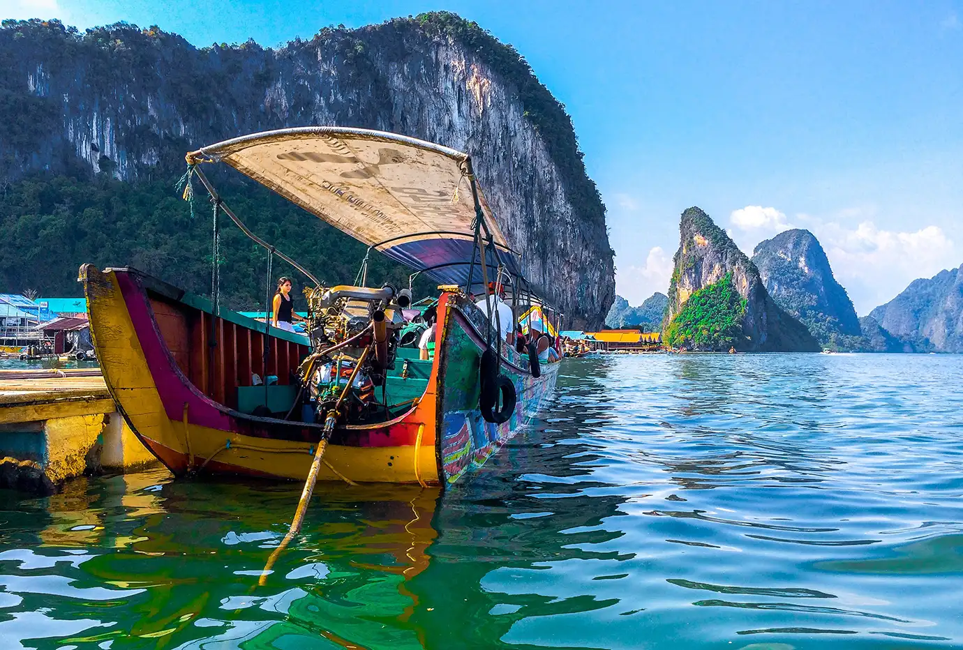 A wide low-angle photograph of a brightly painted wooden longtail boat moored at a floating pier in the emerald waters of Phang Nga Bay, backed by massive limestone karst cliffs in Thailand.