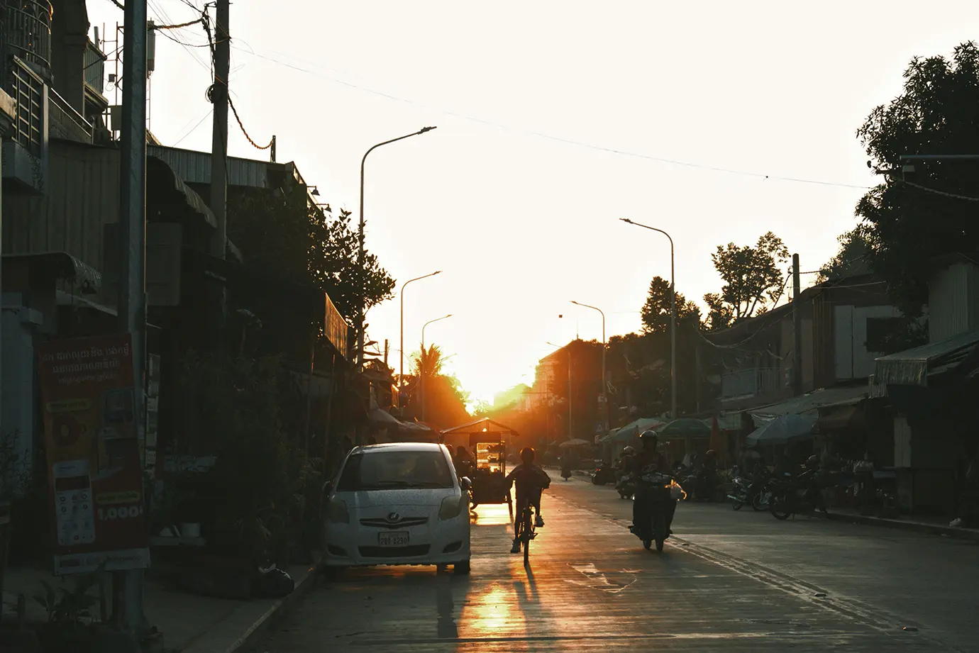 A cyclist riding down a road toward a bright sunset alongside parked cars and motorbikes featured in a travel guide on Is Cambodia Safe.