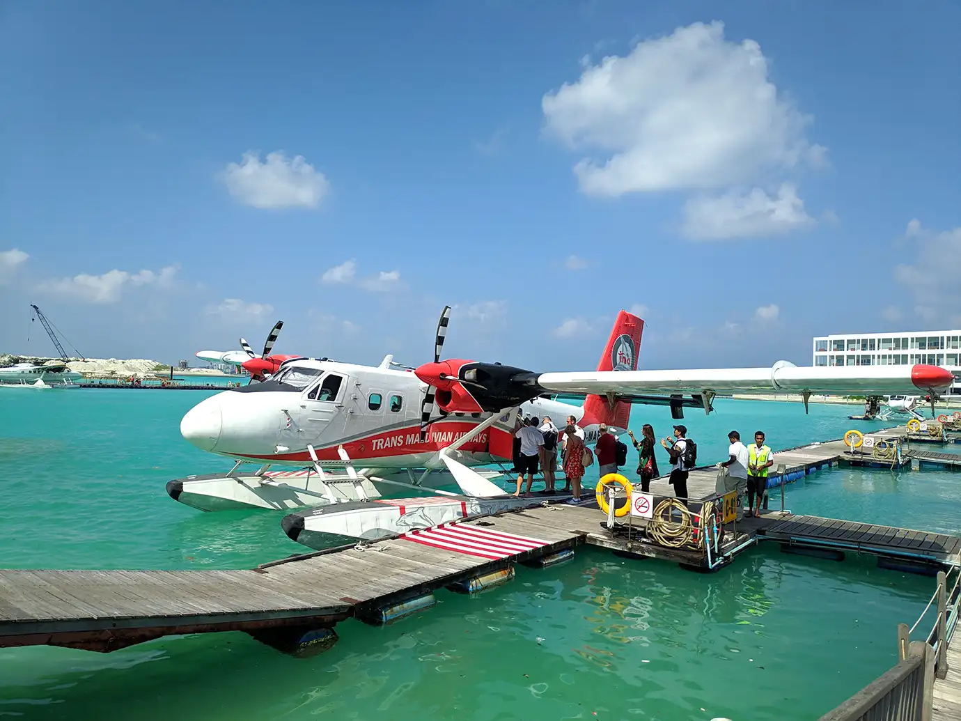 A red and white Trans Maldivian Airways Twin Otter seaplane docked at a wooden floating platform as passengers prepare to board on clear turquoise water in Malé, Maldives.