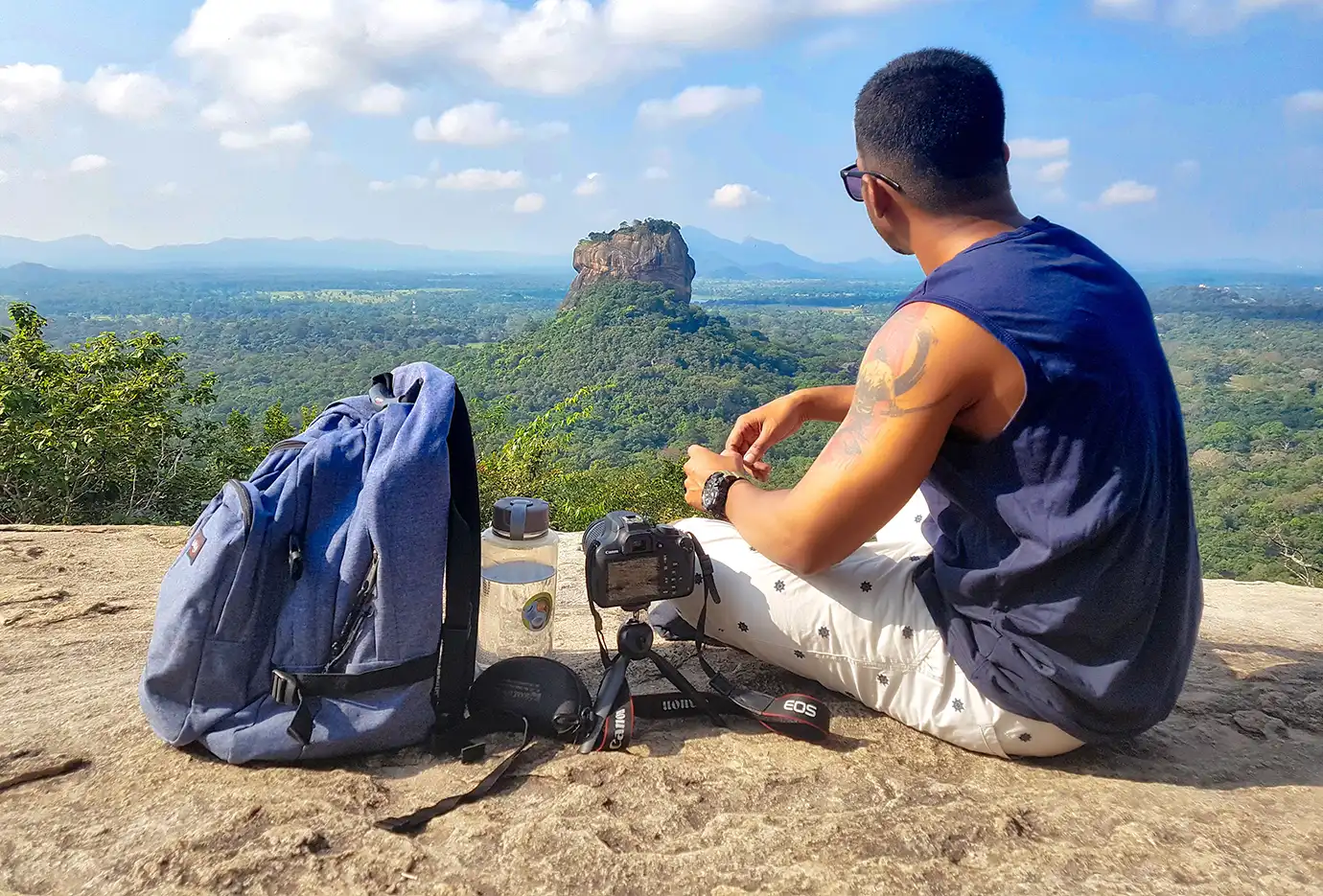 A man sitting on a rocky summit next to a blue backpack and a tripod-mounted camera, gazing across a dense green forest toward Sigiriya Rock under a partly cloudy blue sky.