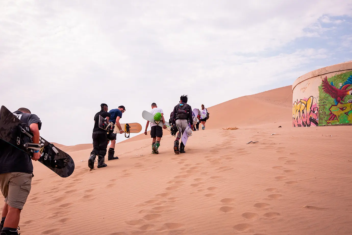 A group of young tourists walking up a high sand dune carrying sandboards during a Dubai Desert Safari.