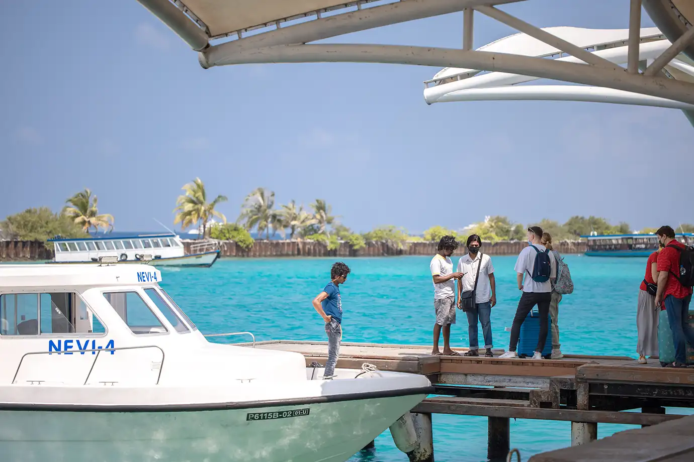 A group of travelers with luggage standing on a wooden jetty next to a white motorboat moored in vibrant turquoise water under a shaded canopy in the Maldives.