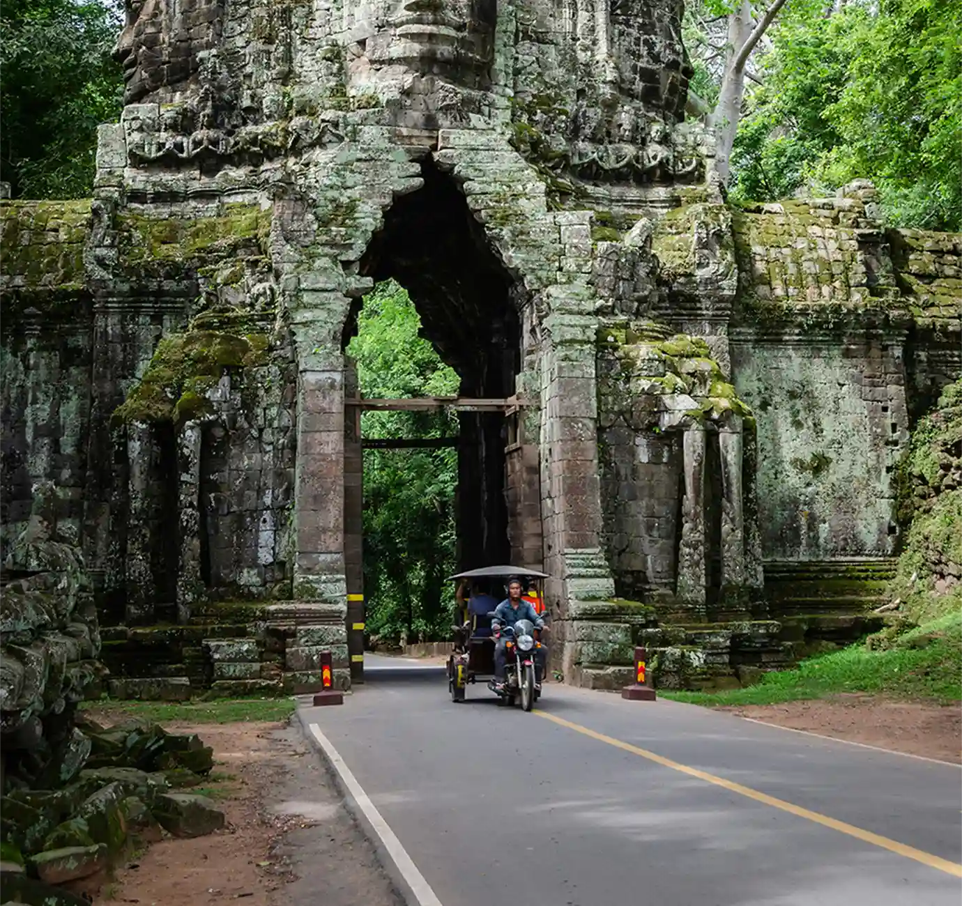 A motorbike tuk-tuk carrying tourists driving through a towering mossy stone archway checkpoint at the Angkor complex featured in a travel guide on Angkor Wat Tickets.