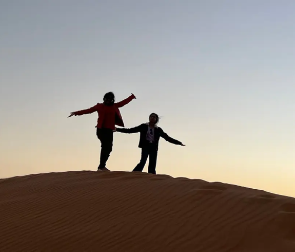 Two travelers silhouetted against a golden twilight sky atop a sand dune featured in an Oman and Maldives Travel Guide.