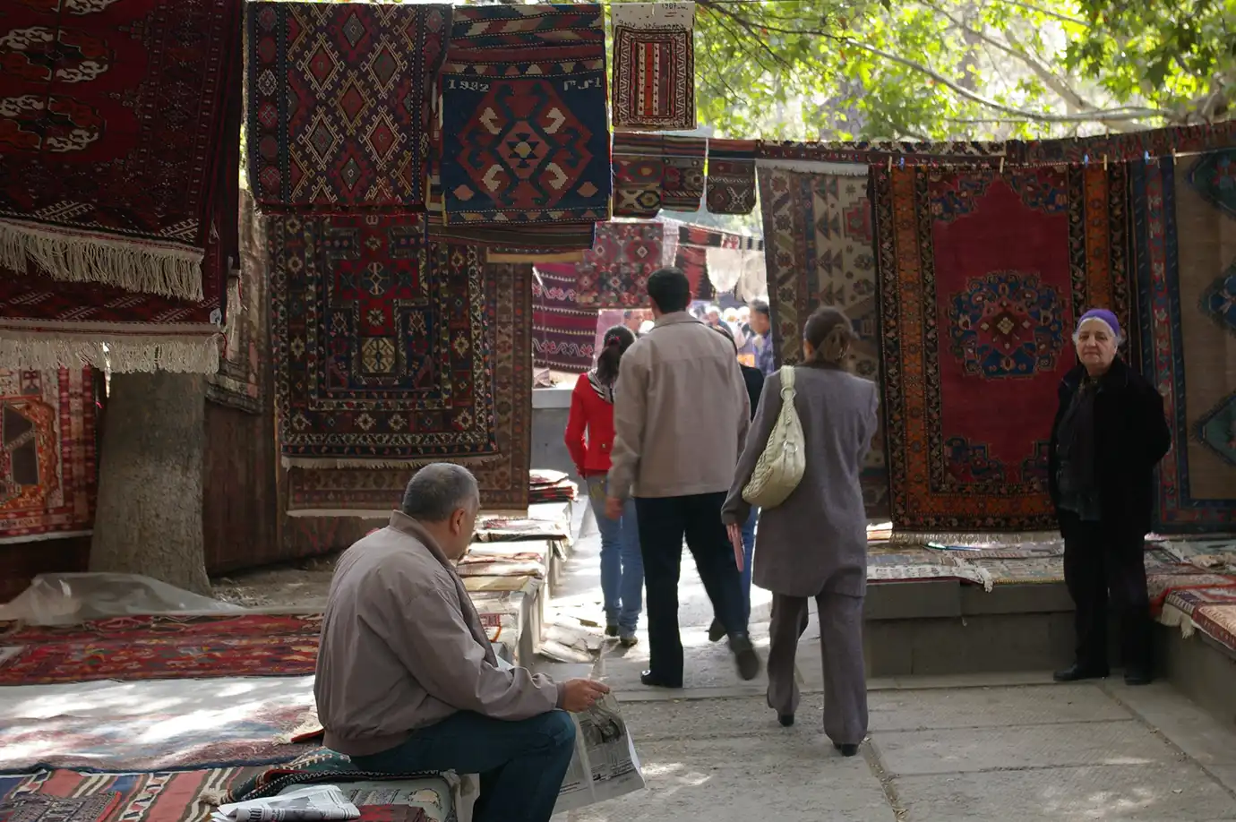 Pedestrians browsing through intricate hand-woven Armenian carpets hanging from stalls at Vernissage Market in Yerevan featured in a travel guide on Best Things to Do in Yerevan Armenia.