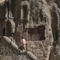 A tourist climbing stone steps past intricate khachkar cross carvings carved into the cliff face at Geghard Monastery featured in a travel guide on 5 Must-See Monasteries in Armenia.