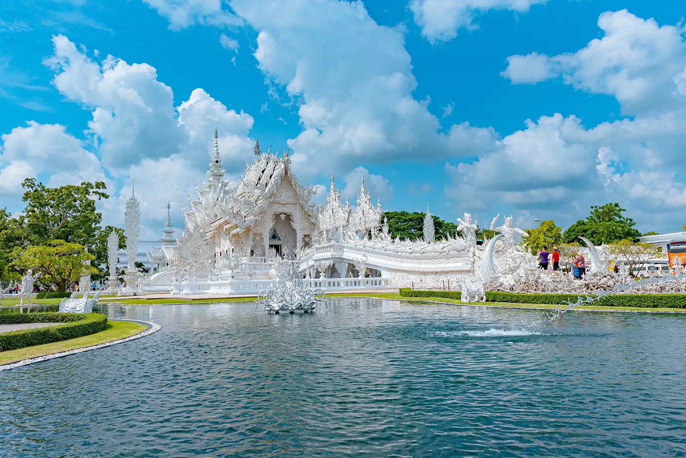 A wide landscape photograph showing the intricate white ornate architecture and bridge of Wat Rong Khun reflected in a large pool of water under a bright cloudy sky in Chiang Rai, Thailand.