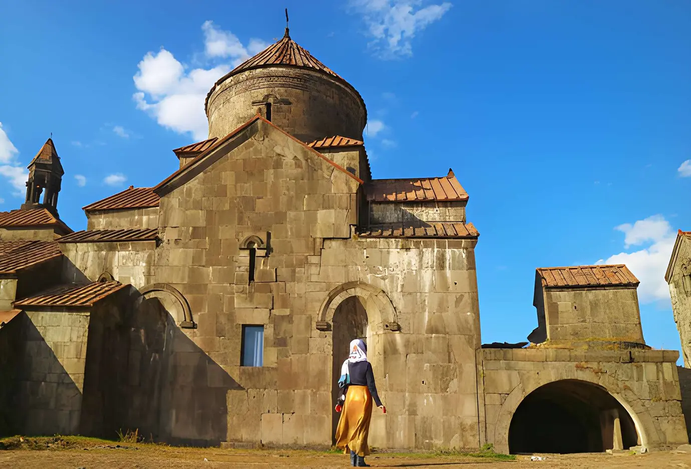 A female tourist in a white headscarf and long golden skirt standing outside the ancient stone walls of Haghpat Monastery featured in a travel guide on What to Wear at Monasteries and Churches.
