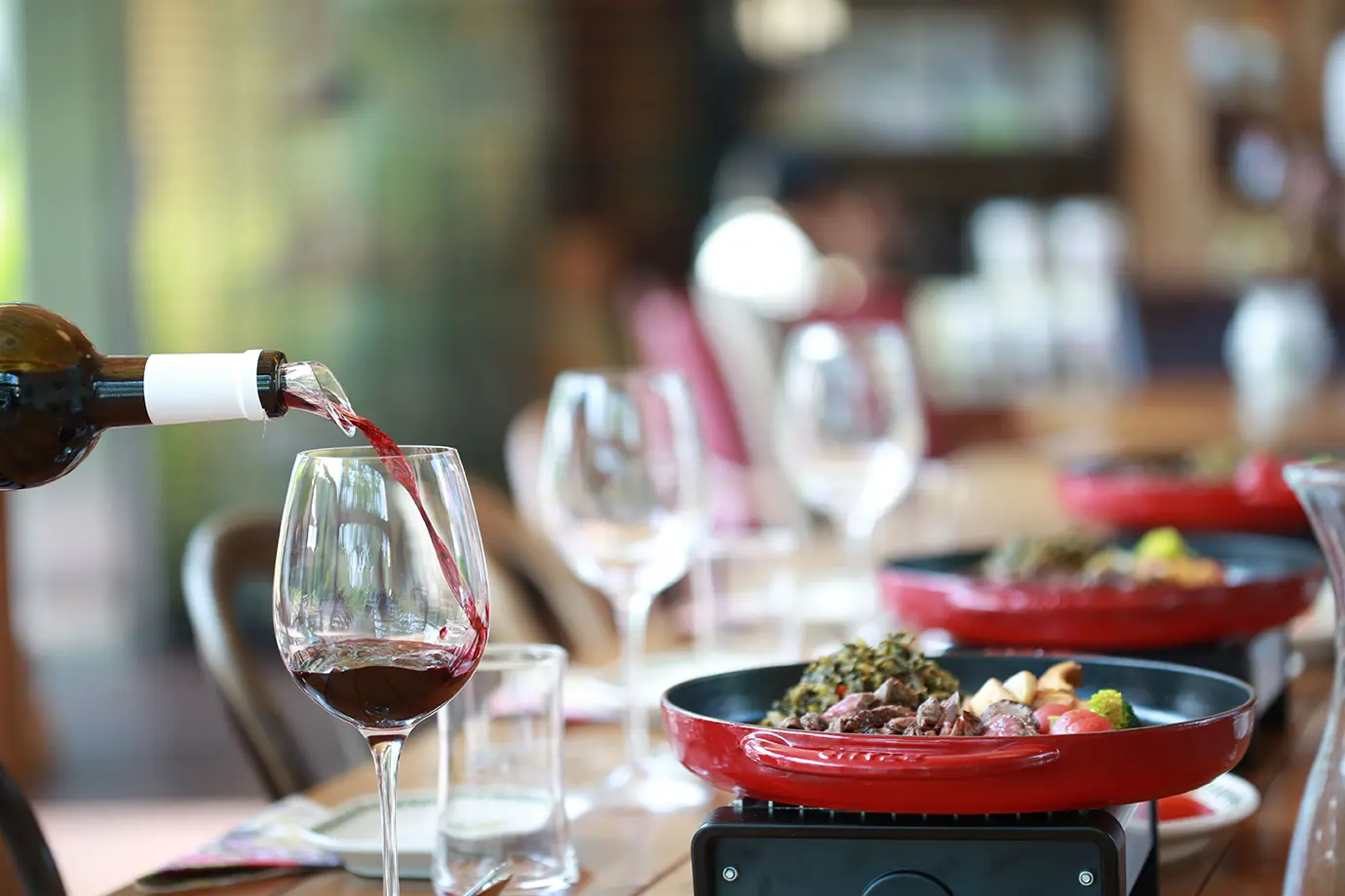 A bottle pouring rich red wine into a clear glass next to hot skillet meals featured in a culinary guide on What Armenian Wine Should You Drink.