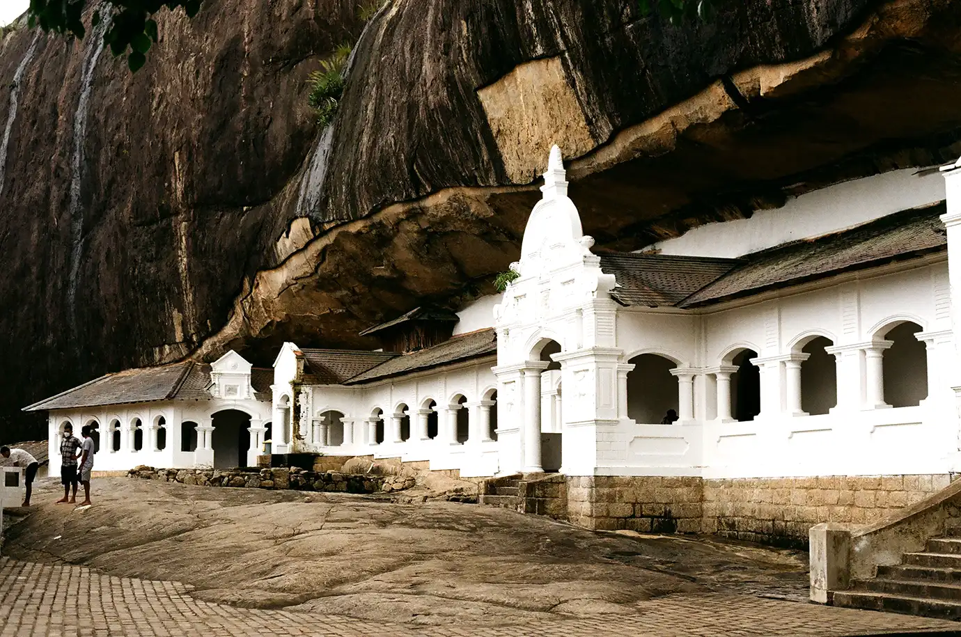 A wide low-angle photograph showing the long, white-arched exterior walls and gabled entryways of the Dambulla Cave Temple nestled beneath a massive, looming granite cliff face in Sri Lanka. 
