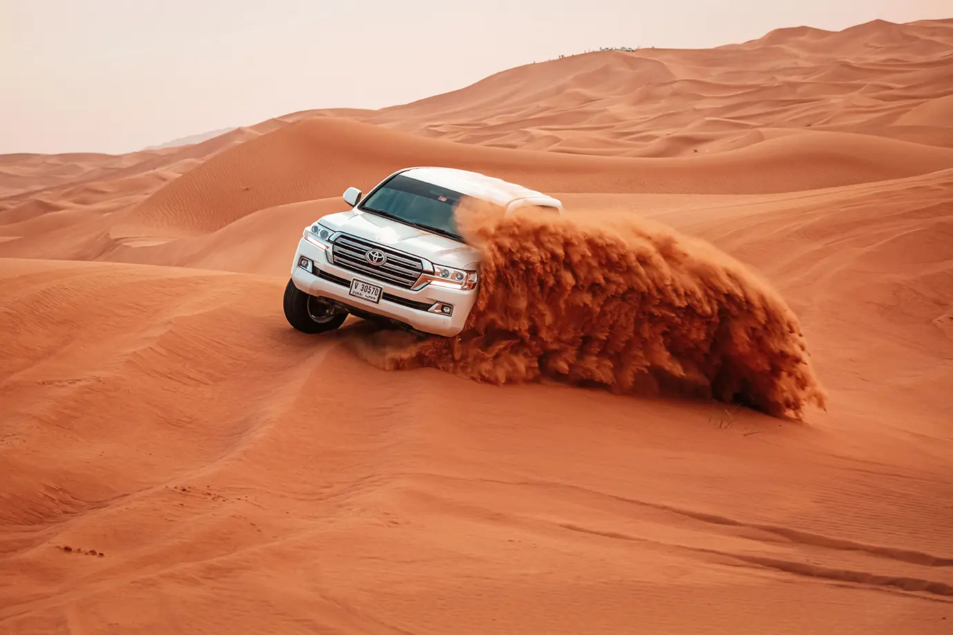 A white SUV drifting down a steep red sand dune, sending up a massive cloud of sand during a Dubai Desert Safari.